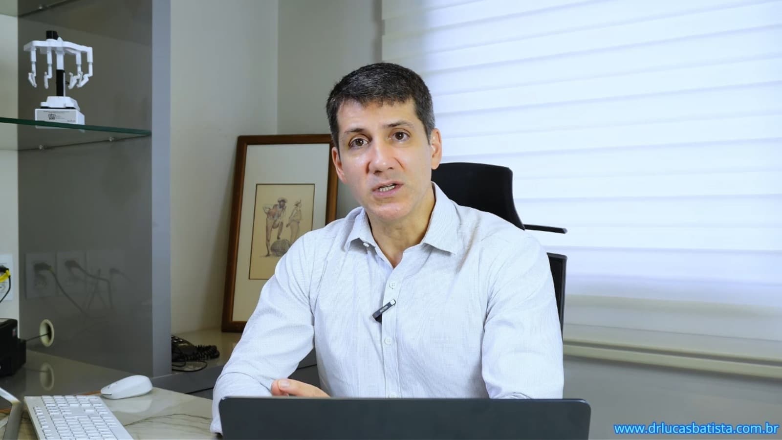 Dr. Lucas Batista talking to camera at his consulting room desk, a surgical robot model on the shelf behind him