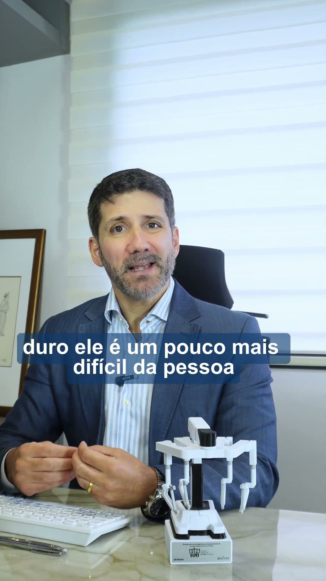 Dr. Lucas Batista speaking at his desk beside a model of a surgical robot, with a burned-in caption