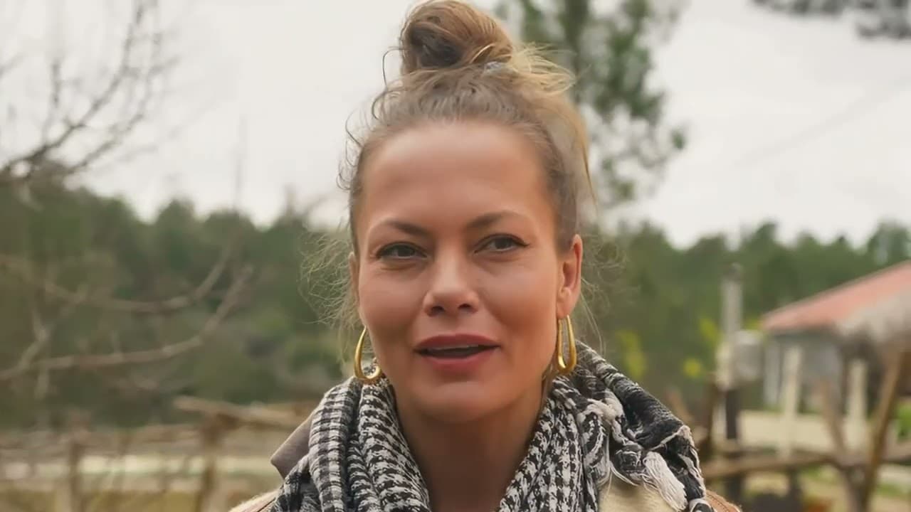 A woman speaking to camera outdoors on a rural property, trees and a shelter behind her