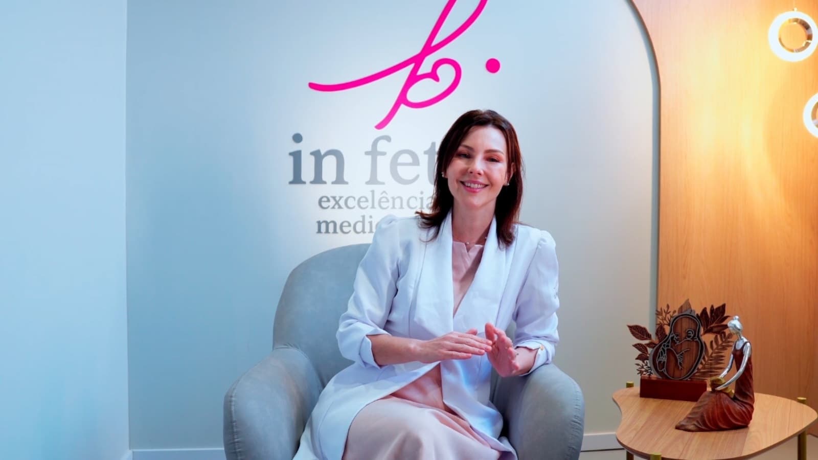 Dr. Patrícia Telló seated in her clinic, smiling at the camera in front of the In Fetus wall, warm light to one side