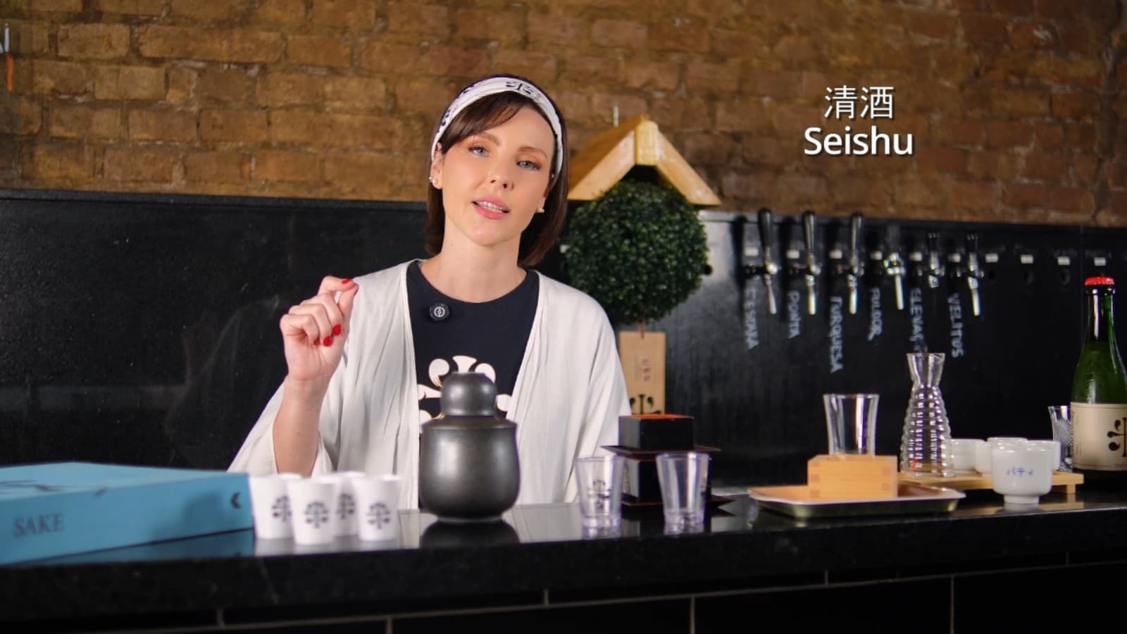 Patty behind a bar with the full sake service laid out in front of her — kettle, cups, carafes and bottles — beside the on-screen title Seishu