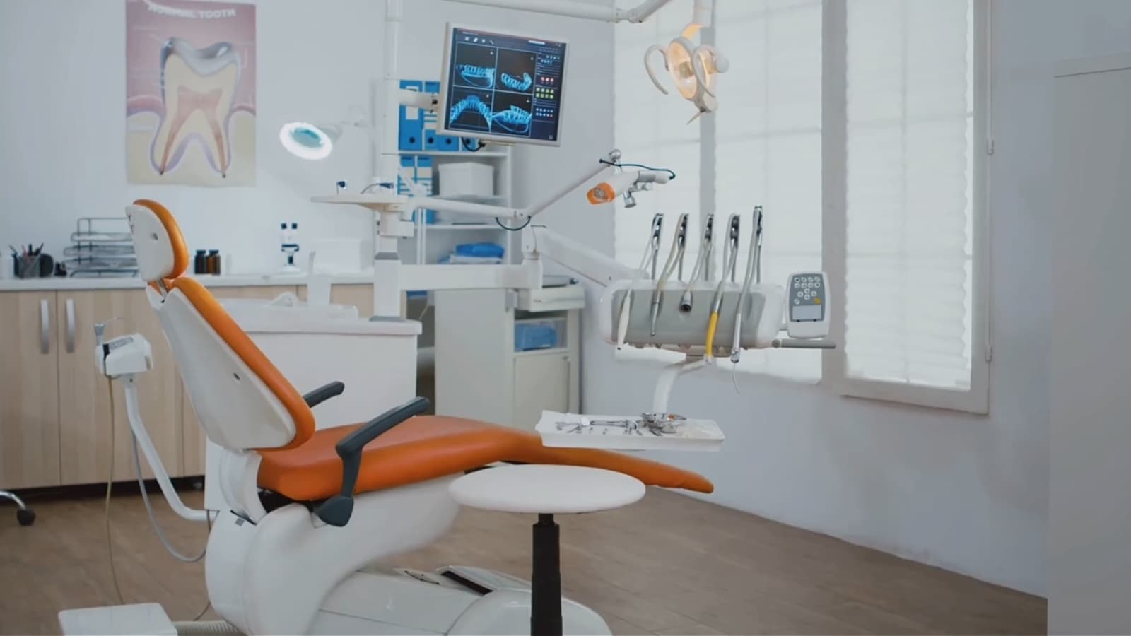 An empty dental surgery, orange treatment chair in the foreground and dental scans on a monitor behind it
